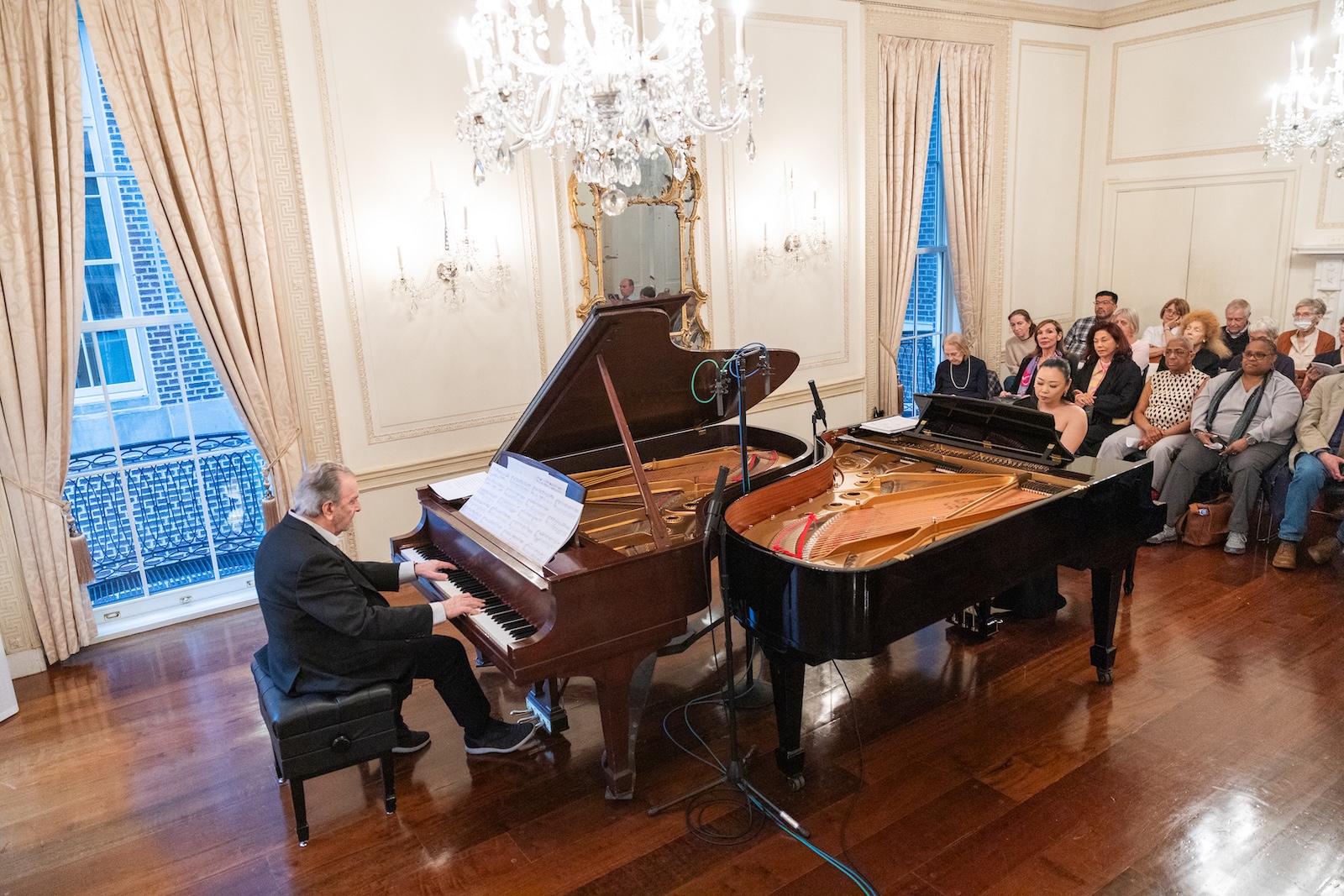 A man and a woman play at two pianos opposite each other in front of an audience in a baroque ballroom.